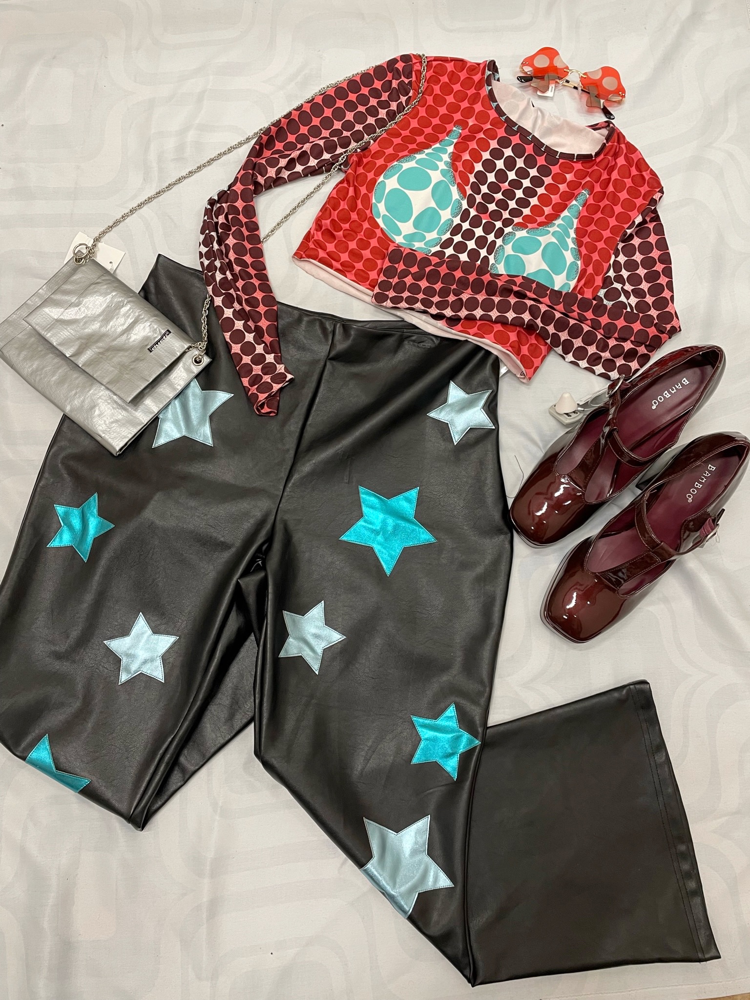 Flat lay of a women's outfit featuring black leather pants with metallic star details, mesh red and blue long sleeve top, red shoes, mushroom sunglasses and a silver chain purse