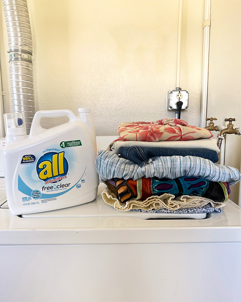 A washing machine with a bottle of All brand detergent and a stack of clothes on top