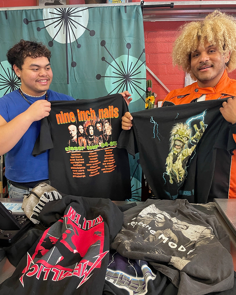 Two Buffalo Exchange Buyers pose behind a counter while holding up tee shirts