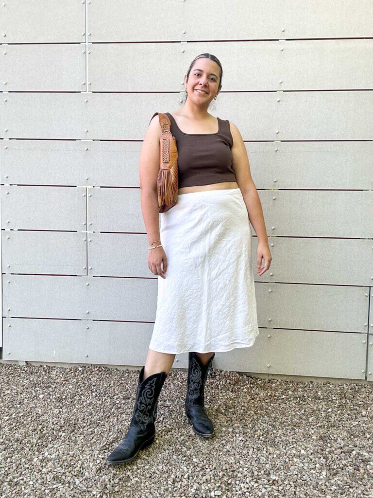 A woman wearing a linen skirt and brown tank top paired with cowboy boots and a leather purse