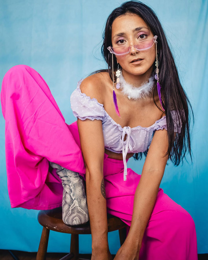 Customer @andreaelenaa_ wearing lilac sunglasses with dangling purple feathers, a lilac off-the-shoulder top, hot pink trousers, and snakeskin boots