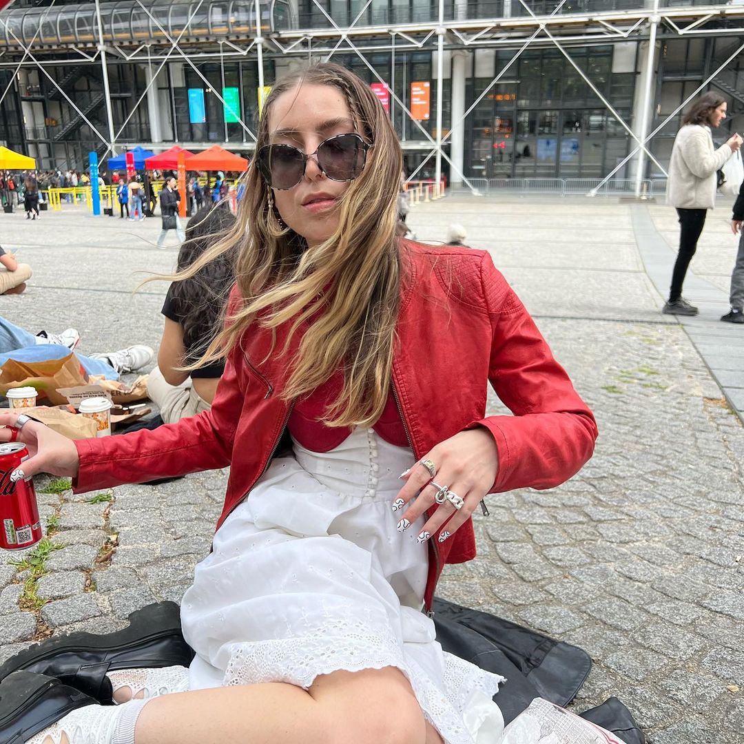 @wantthisnic wearing oversized sunglasses, a red leather moto jacket, and a red dress with a white eyelet corset and skirt.