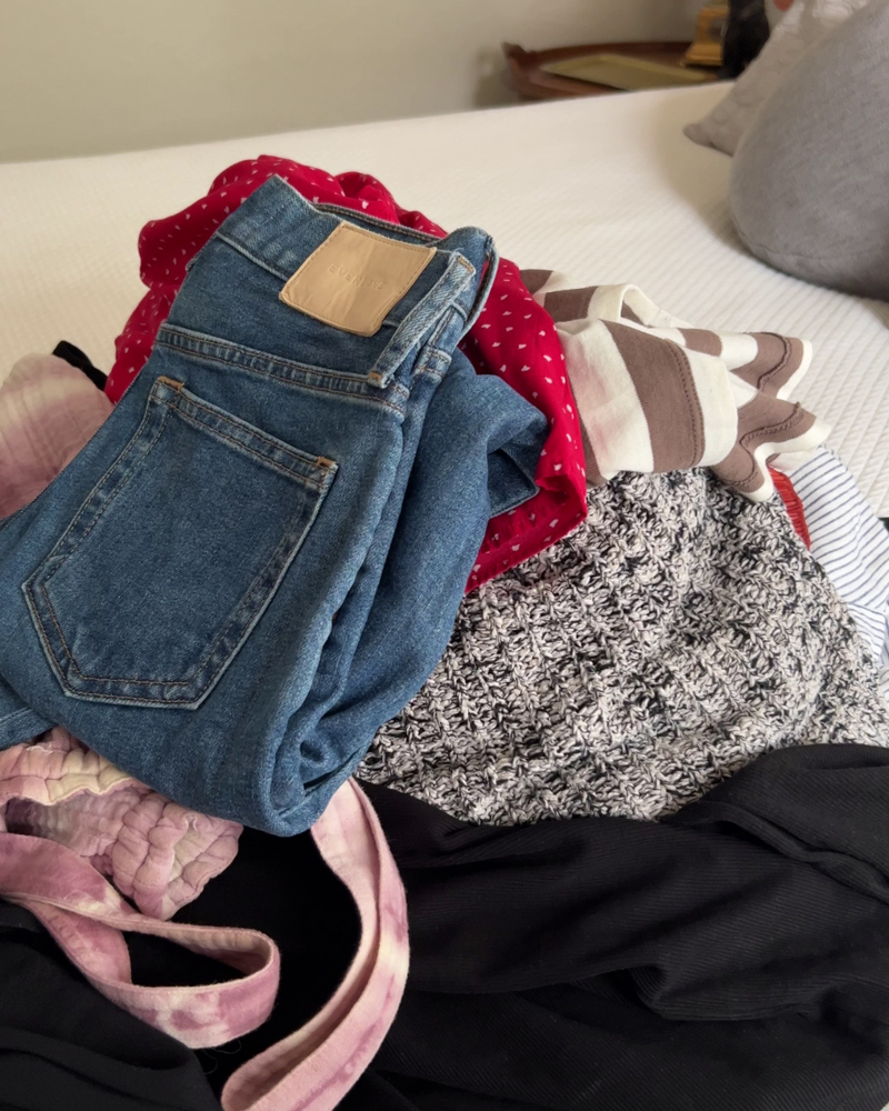 A pile of clothes on a bed, including Everlane jeans, a red polka dot skirt, and a white and grey sweater.