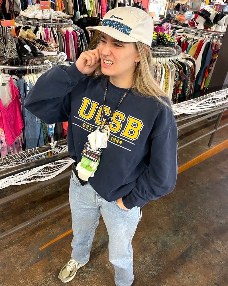 Person wearing UCSB sweatshirt, jeans, sneakers and baseball cap talks on retro cellphone