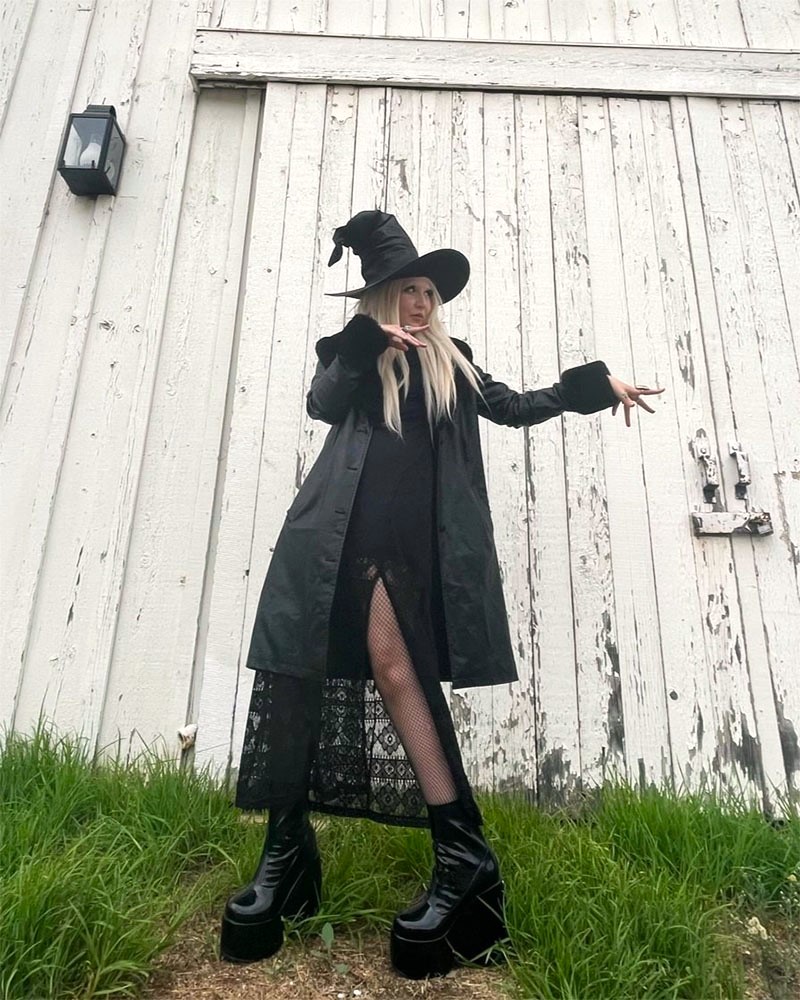 Person dressed in all black witch costume poses in front of barn wall