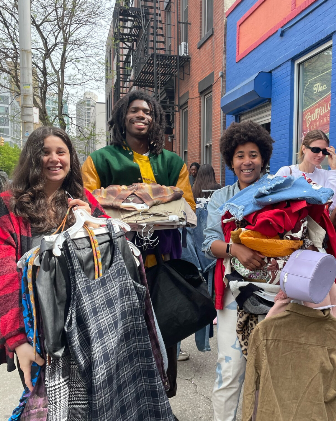 A group of people smiling while holding armfuls of clothing in front of a Buffalo Exchange store
