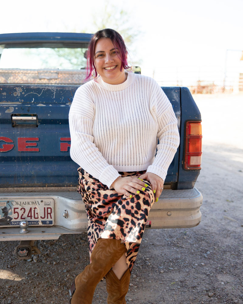 Monique sitting on the back of a truck in a white ribbed sweater, leopard print skirt & cowboy boots