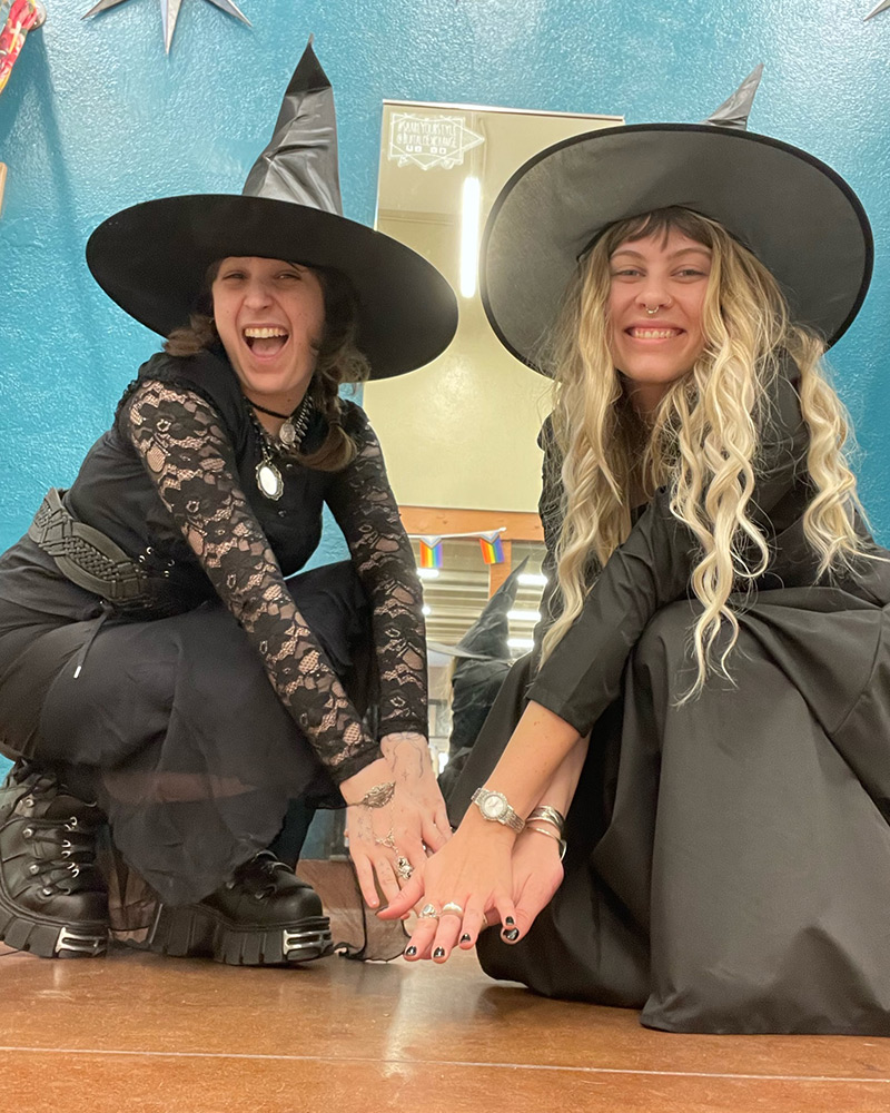 Two women crouch and hold hands wearing witch costumes