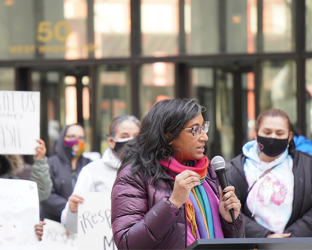 A woman speaking into a microphone in front of a crowd with signs.