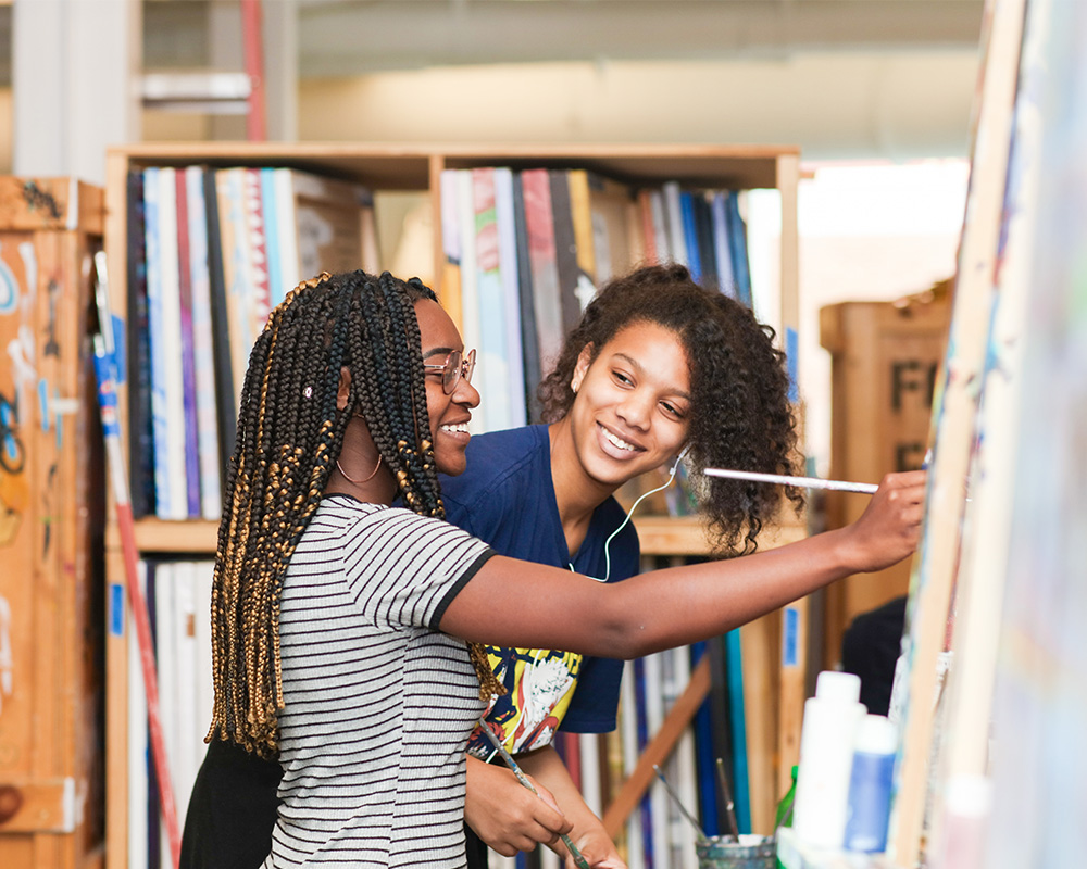 Two teens smiling and painting on a canvas.