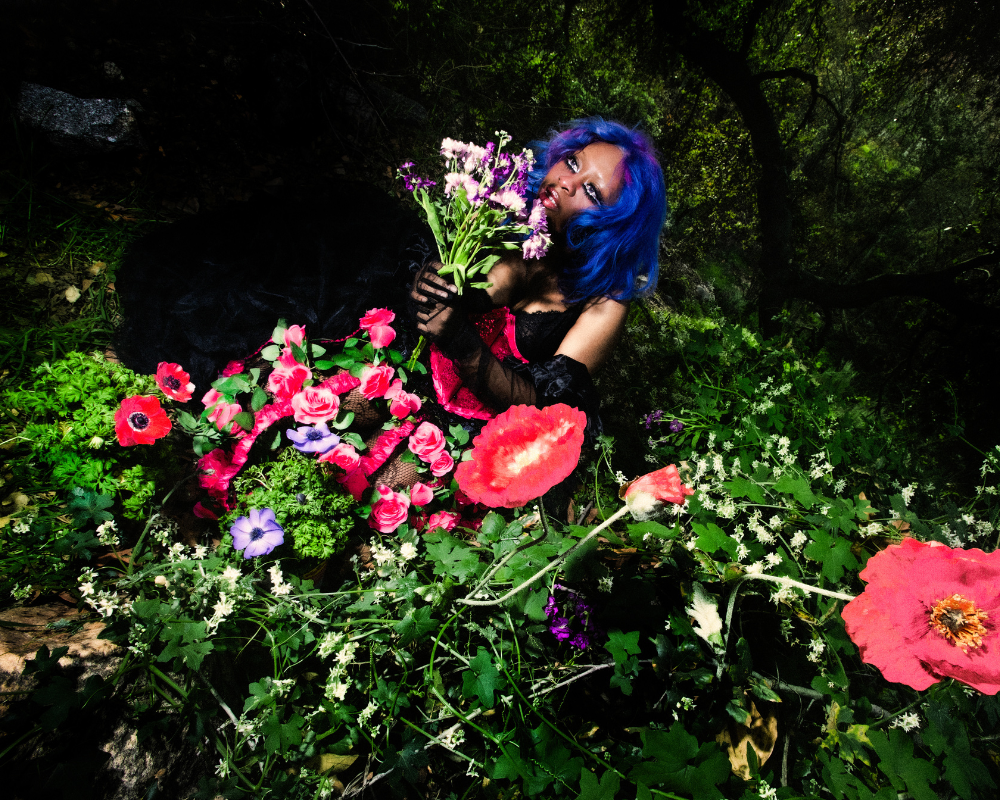 hemlocke springs holds a bouquet while sitting in a bed of flowers against a dark forest background